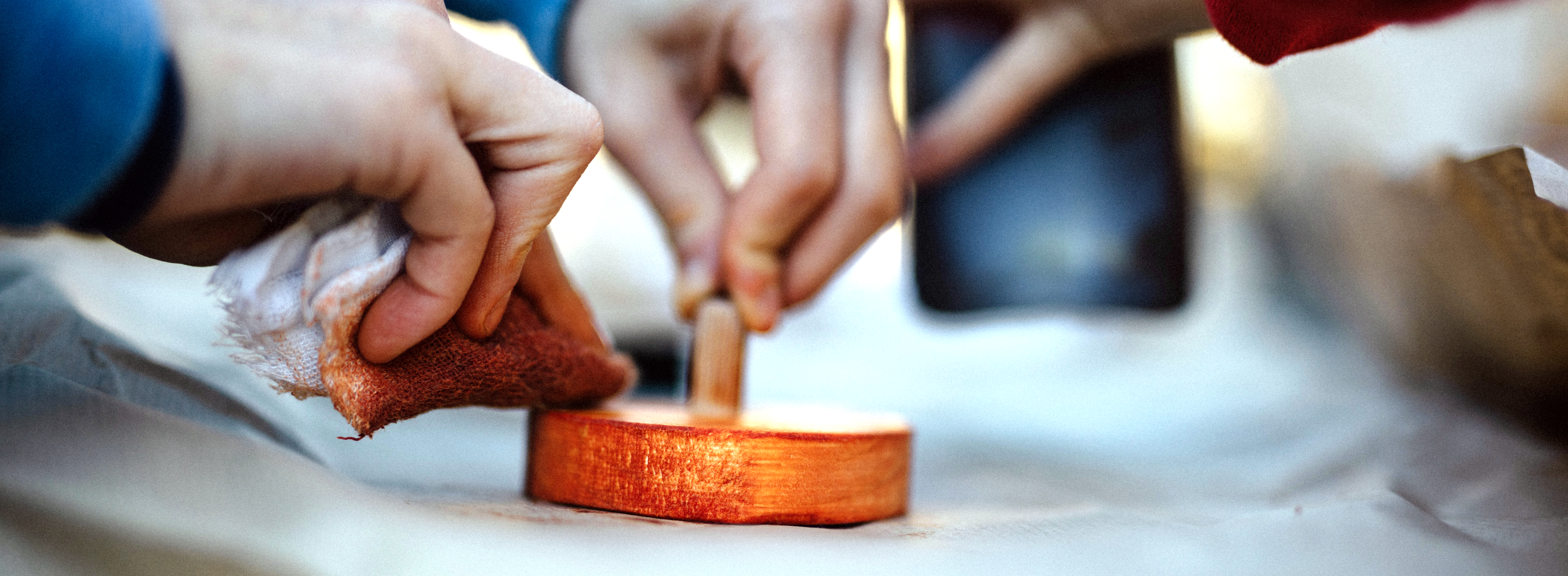 Bild von zwei Händen die mit einem Tuch dunkelorange Farbe auf ein Holzobjekt auftragen